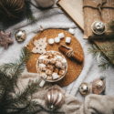 Flat lay of cozy and festive items, including a mug of hot chocolate and marshmallows, cinnamon sticks, pine leaves, Christmas ornaments, a book, and a gift.