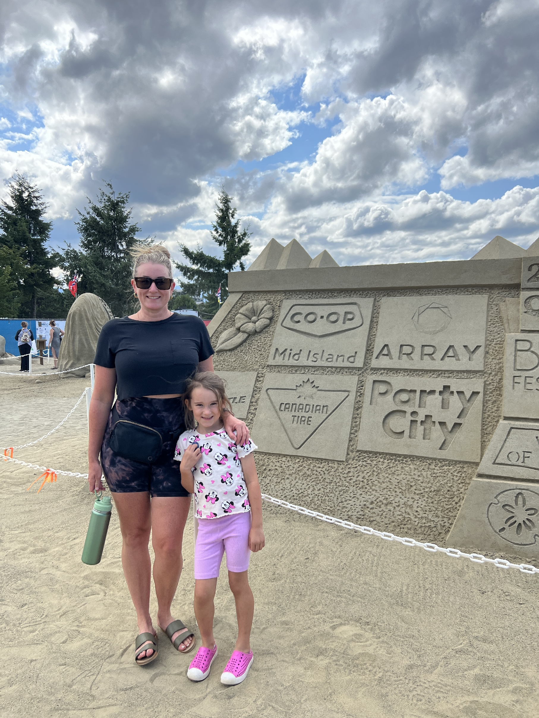Kristina Crowson and child pose together at a community event. Behind them is a detailed sand sculpture featuring logos of companies sponsoring the event, including Array.