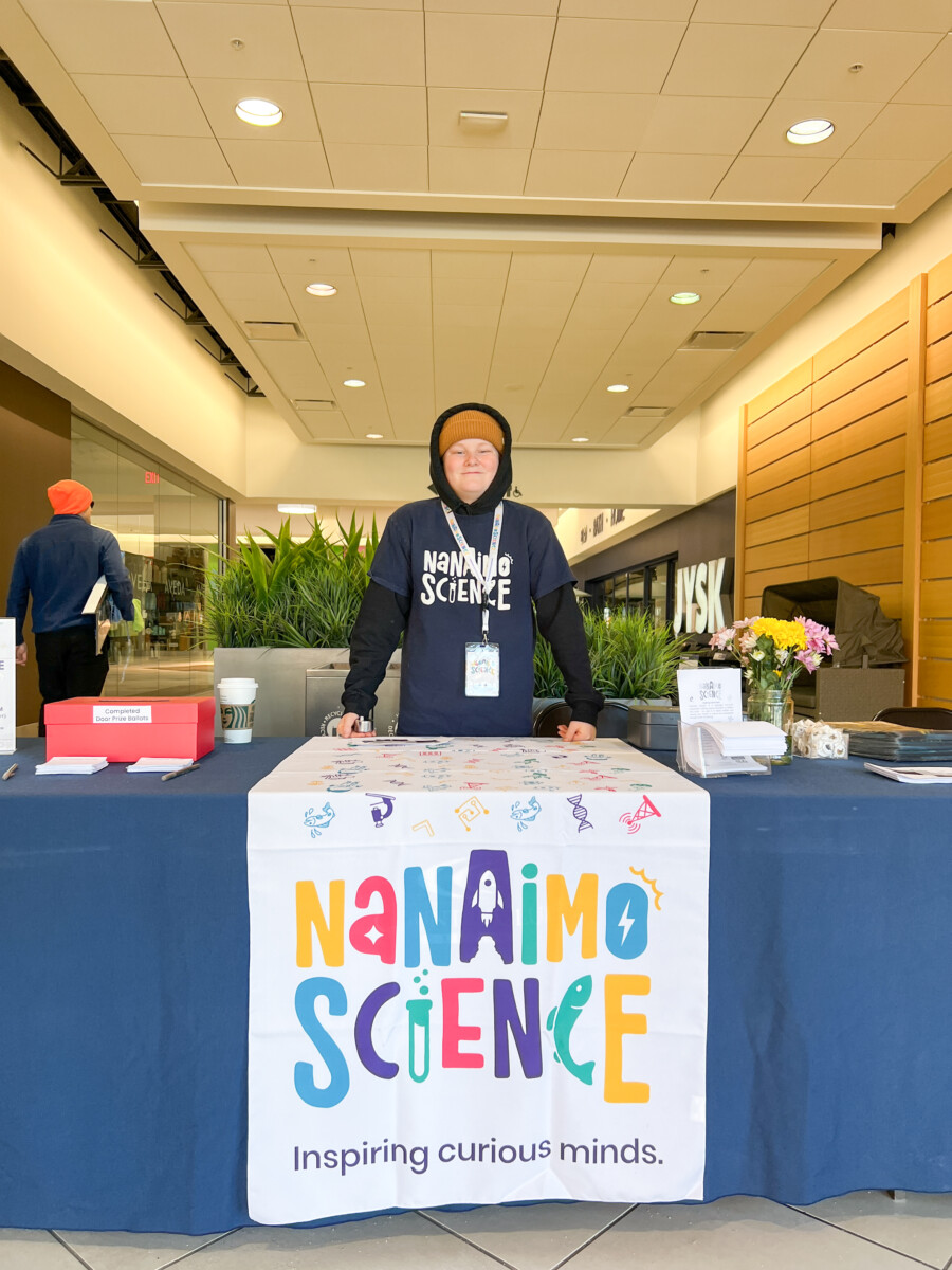 A person stands behind a booth representing Nanaimo Science in an indoor setting, wearing a hoodie with 'Nanaimo Science' printed on it.