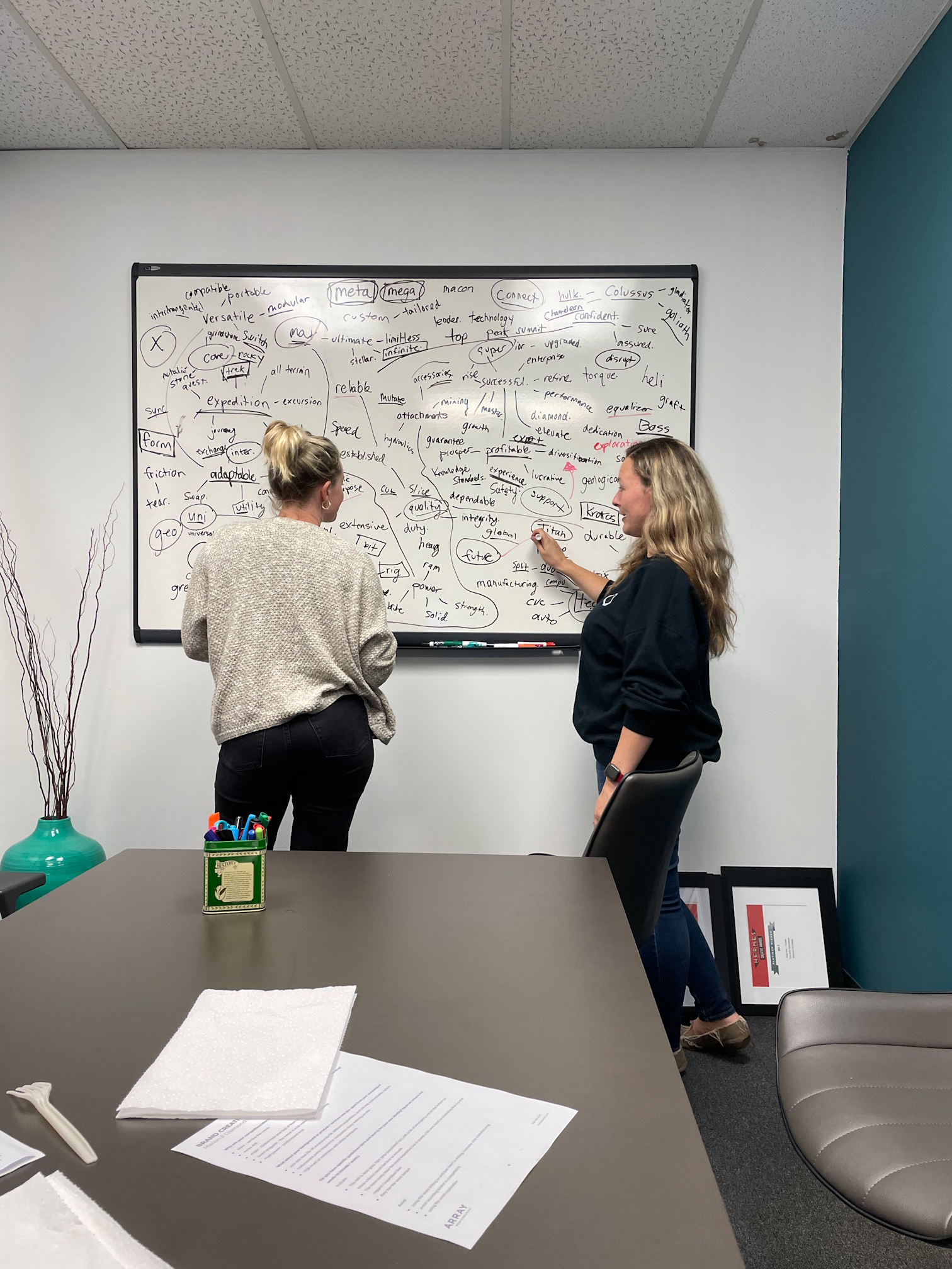 Two women writing on a white board in an office meeting room.