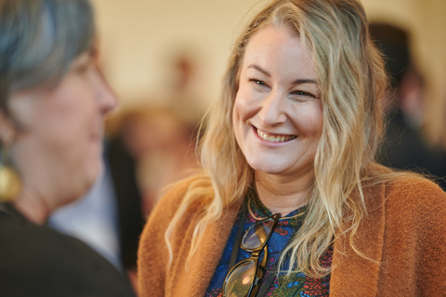 A close-up of a smiling blonde woman, wearing a light brown coat over a colorful patterned shirt. She is in conversation with someone slightly out of focus in the foreground, with a warm and friendly expression, captured in a bright, indoor setting.