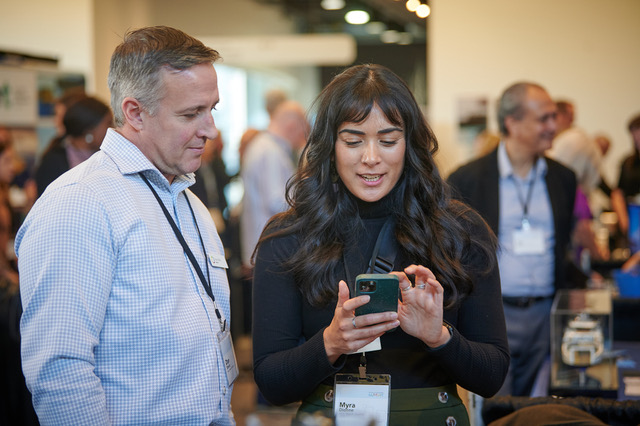 A candid photo of two conference attendees, a man in a light blue shirt and a woman with dark hair and a black top. The woman is showing something on her smartphone while the man observes, both wearing event badges in a busy networking space.