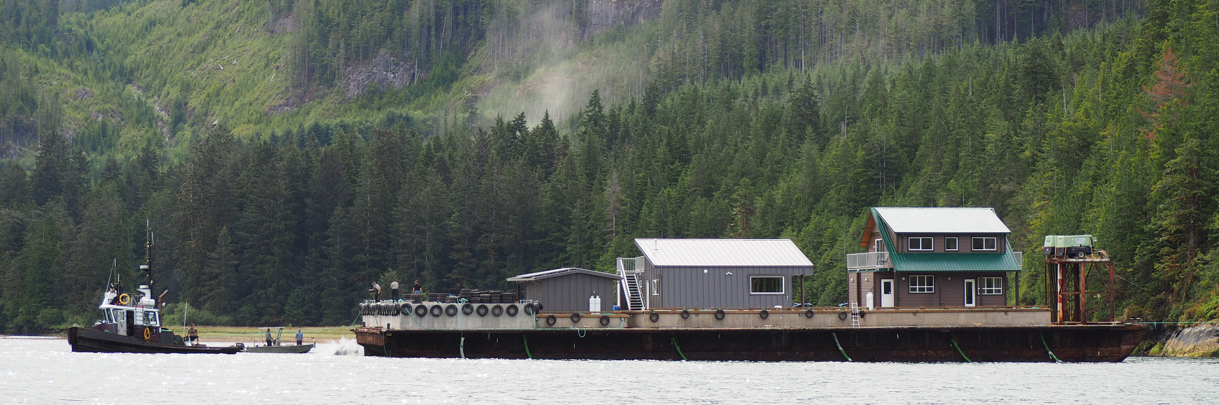 Floating dock with homes and small boats on the water. There is a lush evergreen forest in the background.